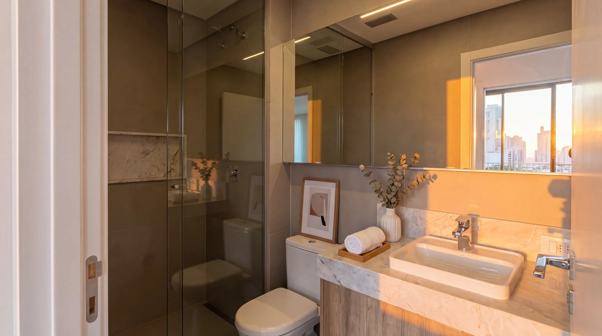 Banheiro social com box de vidro, bancada em mármore claro com cuba retangular de louça, gabinete em madeira clara, espelho amplo com iluminação LED linear superior, revestimento efeito pedra na parede e janela basculante com luz do fim de tarde