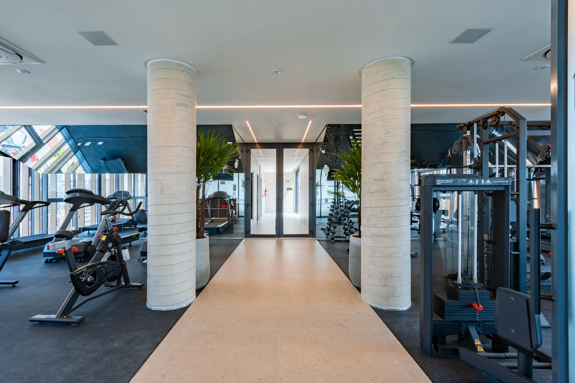 Academia com bicicletas ergométricas, aparelhos de musculação Movement e colunas em concreto aparente com iluminação LED indireta