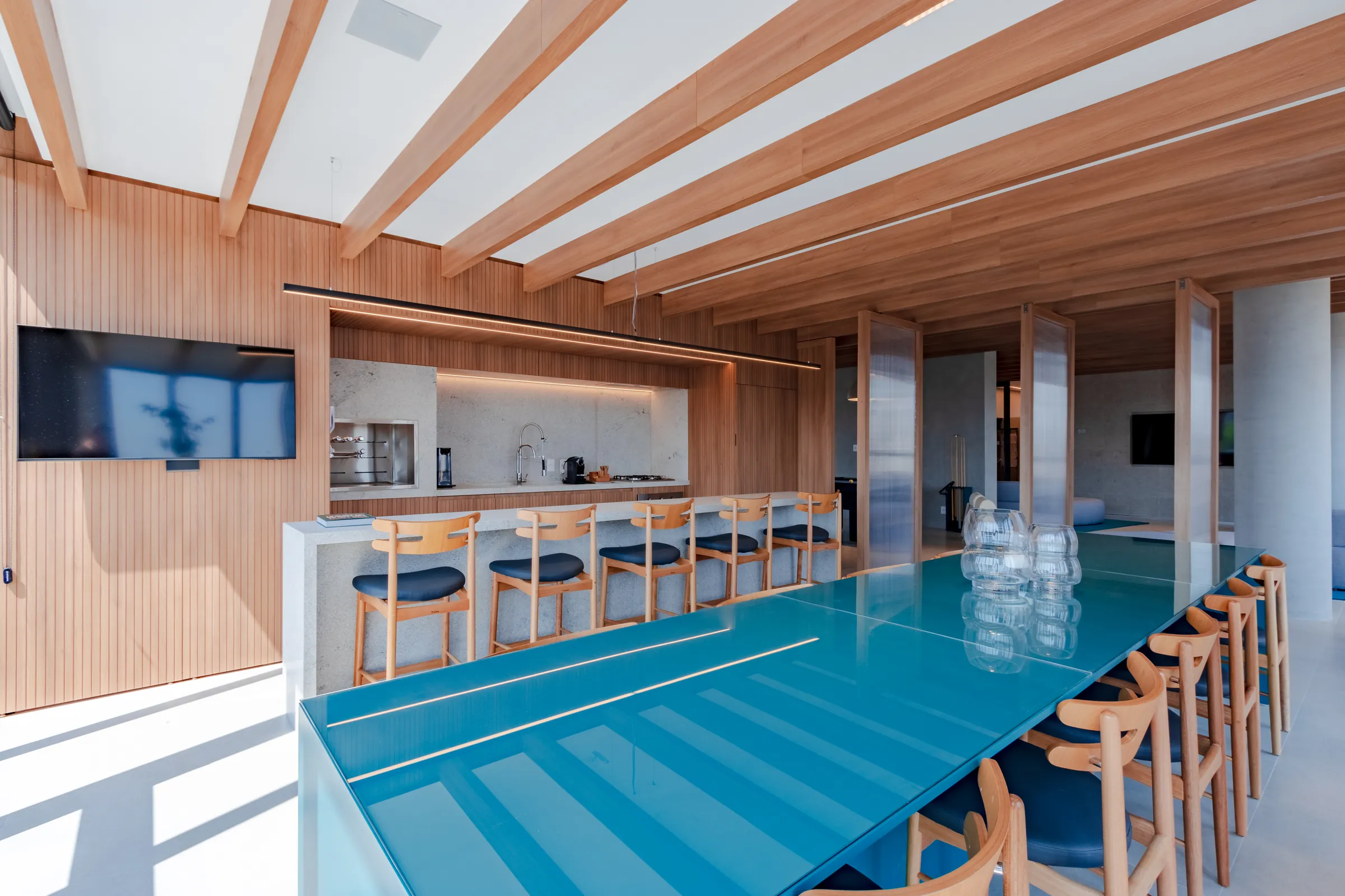 Clubhouse com mesa longa de vidro azul-turquesa, cadeiras de madeira, balcão com bancos, TV e teto em vigas de madeira clara