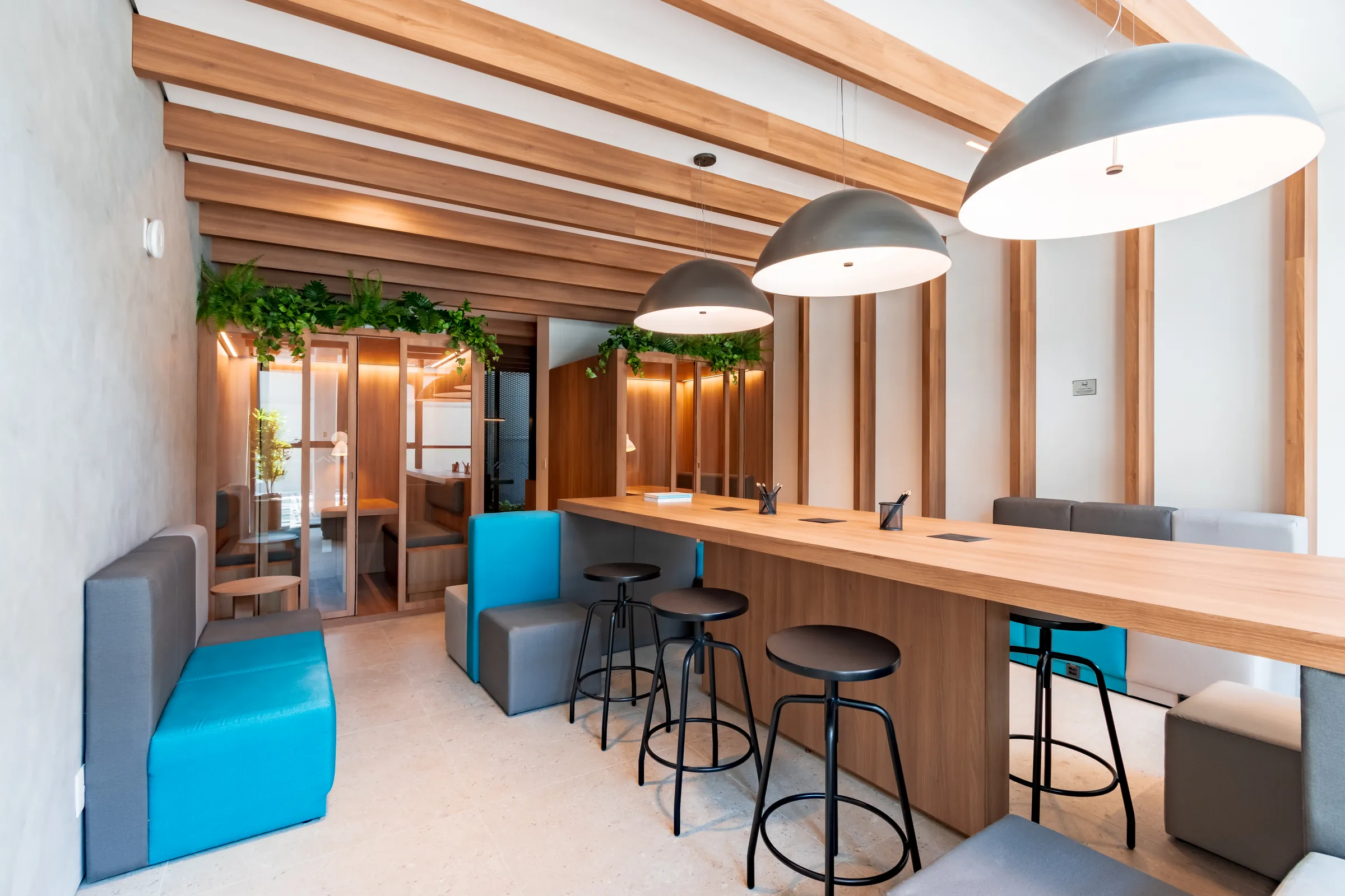 Coworking do Vaz Batel com balcão longo de madeira natural, banquetas pretas, bancos estofados em cinza e azul, luminárias pendentes grandes e cabines de reunião ao fundo