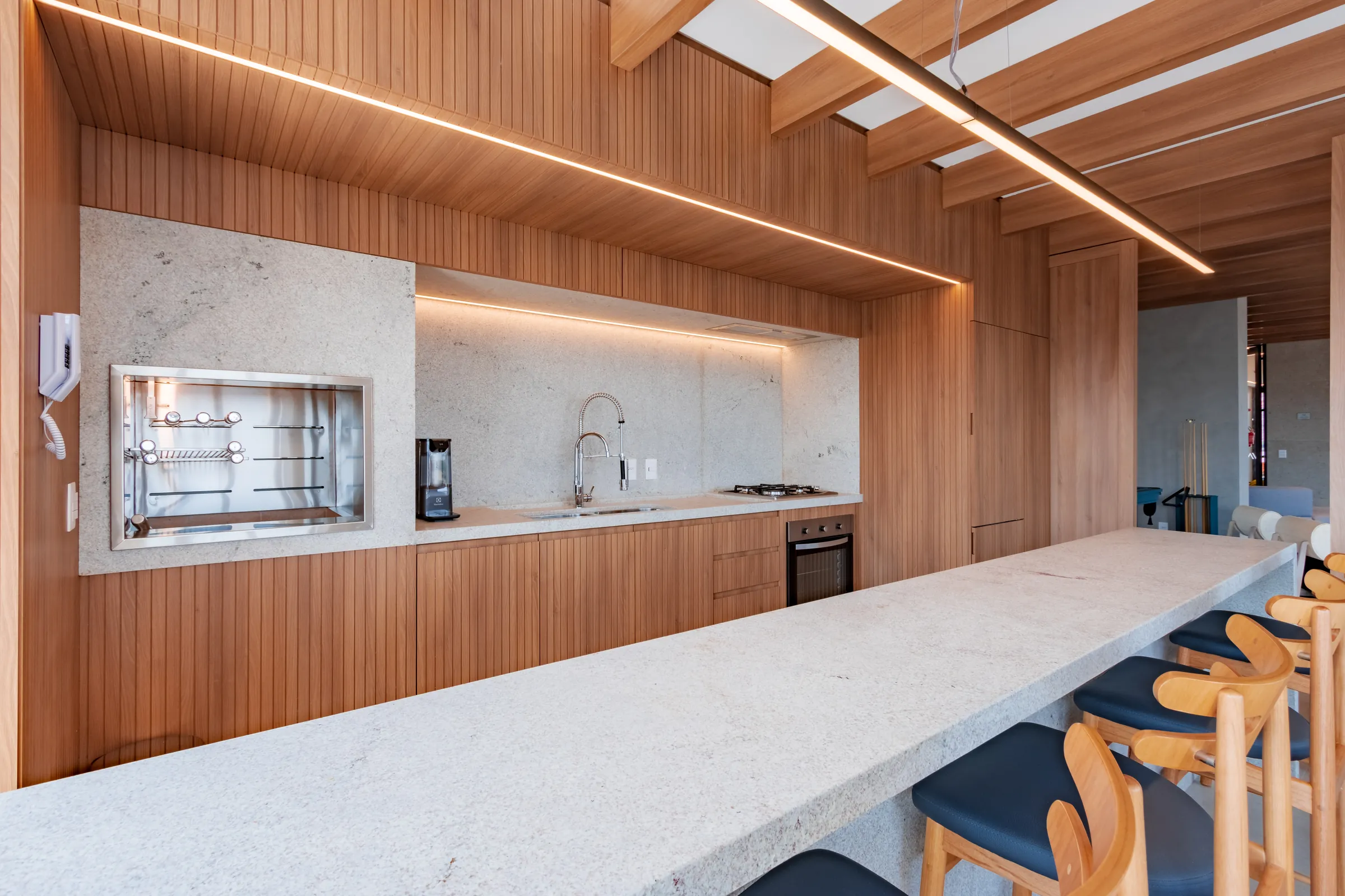 Espaço gourmet com churrasqueira elétrica com espetos rotativos, bancada de mármore, armários em madeira e iluminação LED linear