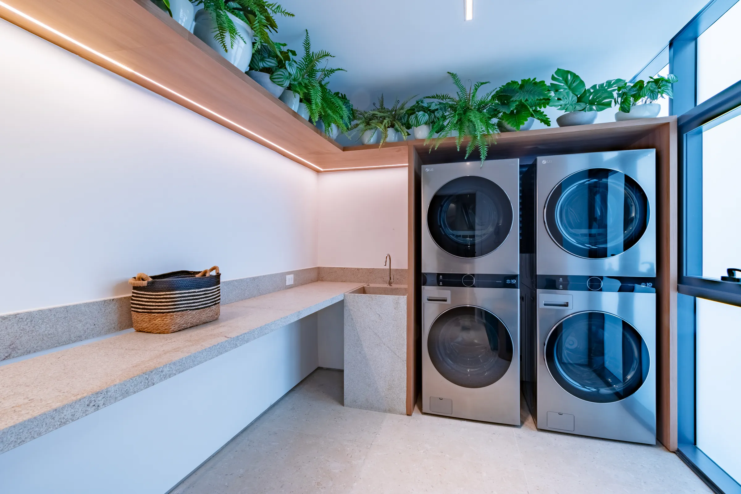 Lavanderia profissional com quatro máquinas LG empilhadas verticalmente em inox, bancada em granito, plantas ornamentais no alto com iluminação LED e janelas laterais