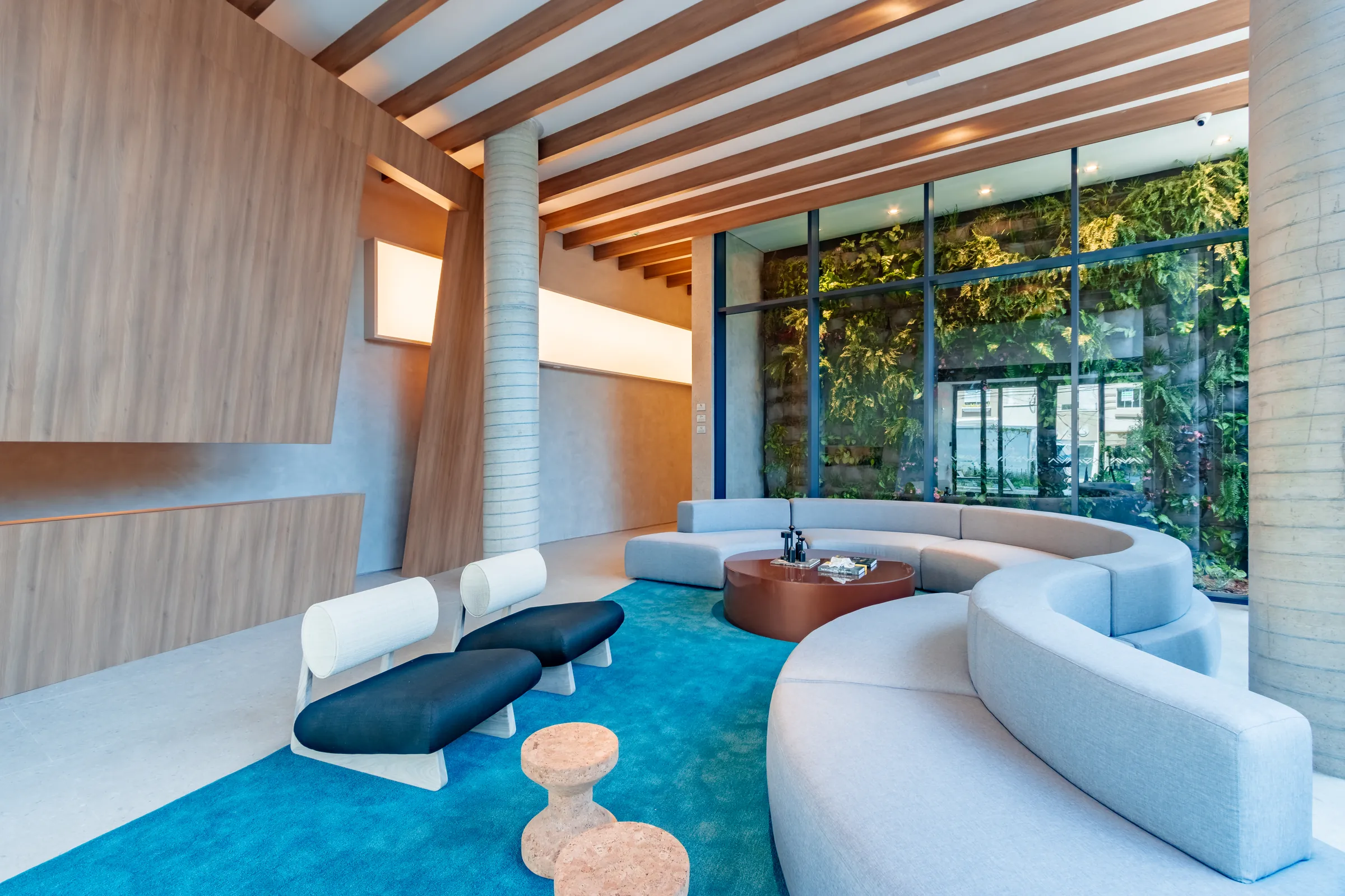 Lobby do Vaz Batel com parede viva de samambaias ao fundo, sofá curvo claro, tapete azul, mesa baixa de madeira e teto em vigas de madeira