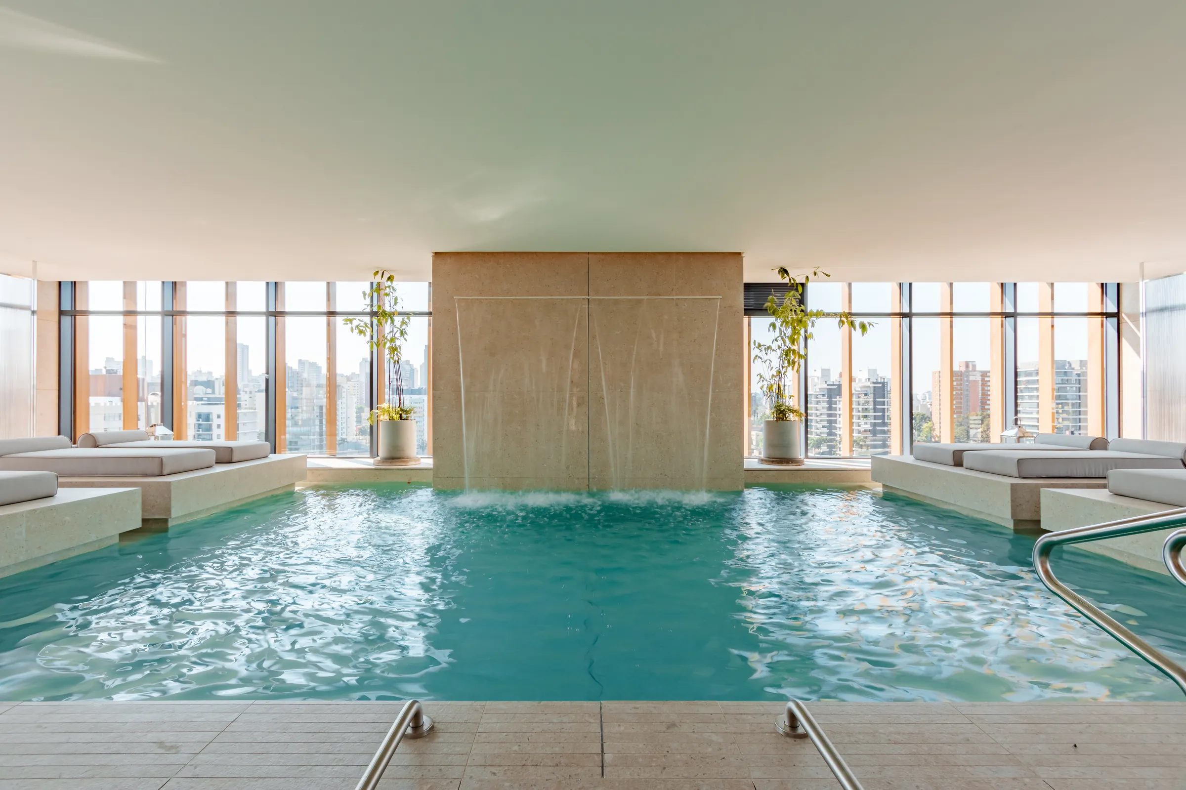 Piscina aquecida do Vaz Batel com cascata central em pedra natural, chaises de concreto integradas nas laterais e janelas do piso ao teto com vista da cidade ao entardecer