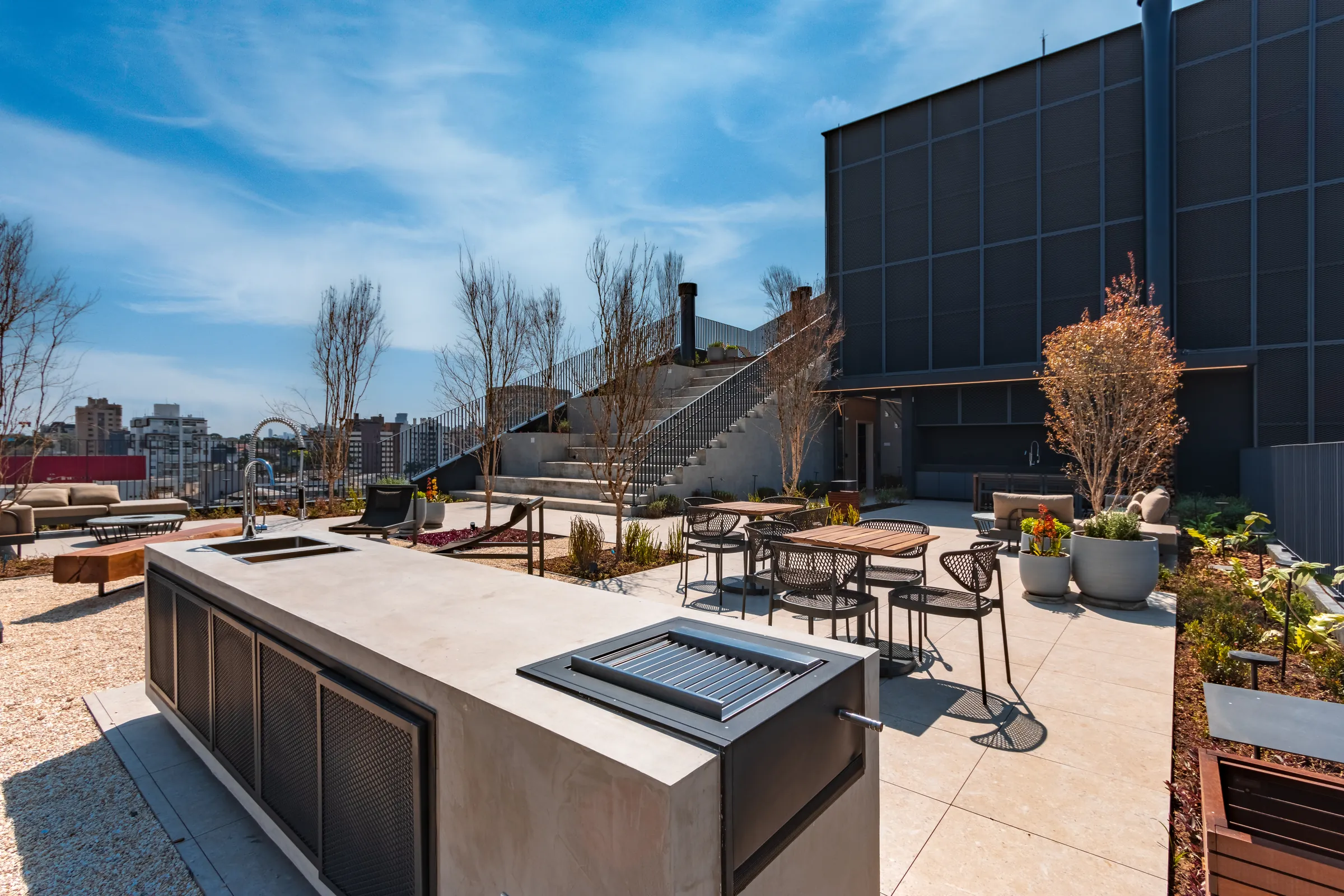 Rooftop externo do Vaz Batel durante o dia com churrasqueira de concreto e aço, mesas altas com cadeiras de metal, paisagismo seco e vista do skyline do Batel ao fundo