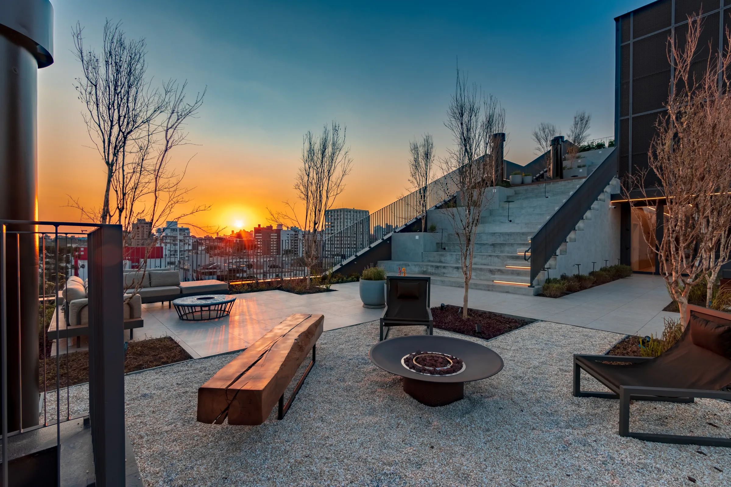 Lounge do rooftop do Vaz Batel com sofás em tom areia e almofadas, mesa baixa redonda em pedra, vasos de plantas e skyline do Batel ao fundo durante o dia