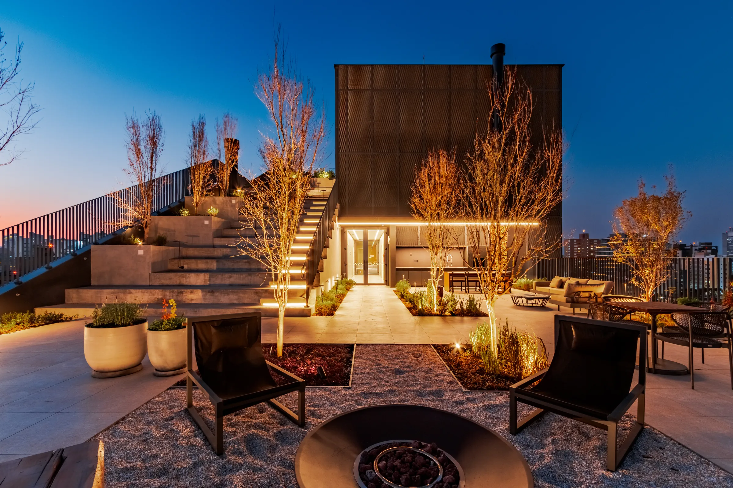 Rooftop do Vaz Batel ao pôr do sol com fire pit aberto, banco em madeira maciça, espreguiçadeiras, paisagismo com árvores e escadaria iluminada