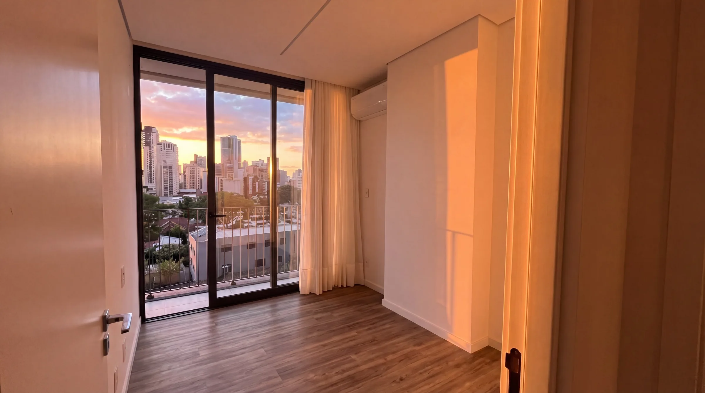 Segundo quarto vazio com piso de madeira natural, paredes brancas, porta-janela do piso ao teto abrindo para sacada com vista do Batel ao entardecer, ar-condicionado split instalado e cortinas leves em tom marfim